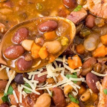 A close-up of bean and vegetable soup with shredded cheese and herbs, a spoon resting in the bowl.