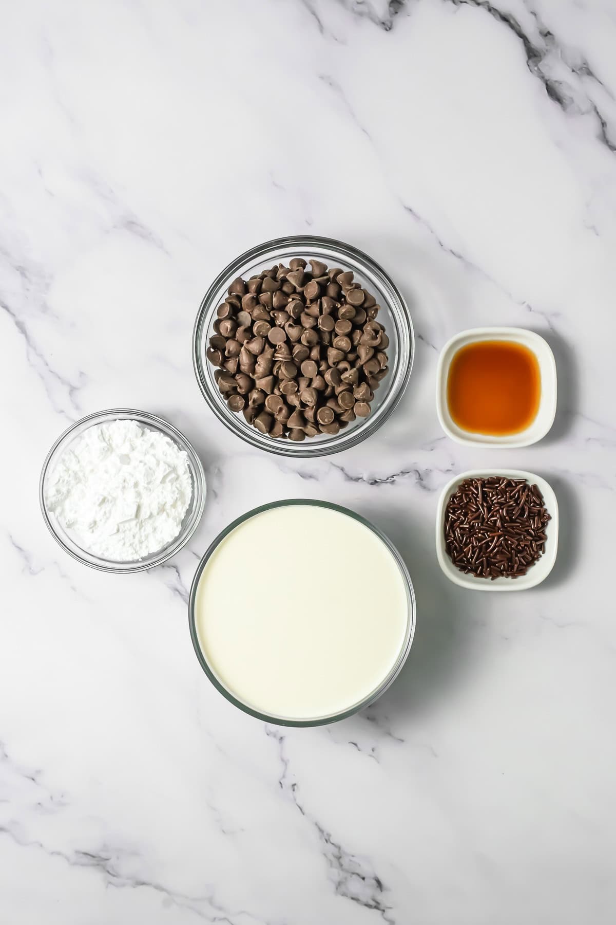 Bowls of chocolate chips, powdered sugar, cream, chocolate sprinkles, and vanilla on a marble surface&mdash;perfect ingredients for creating a rich chocolate mousse.