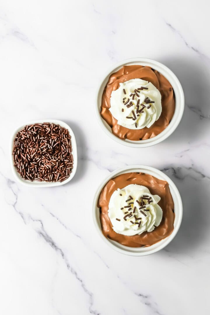 Two bowls of chocolate mousse pudding topped with whipped cream and chocolate sprinkles, beside a dish of sprinkles.