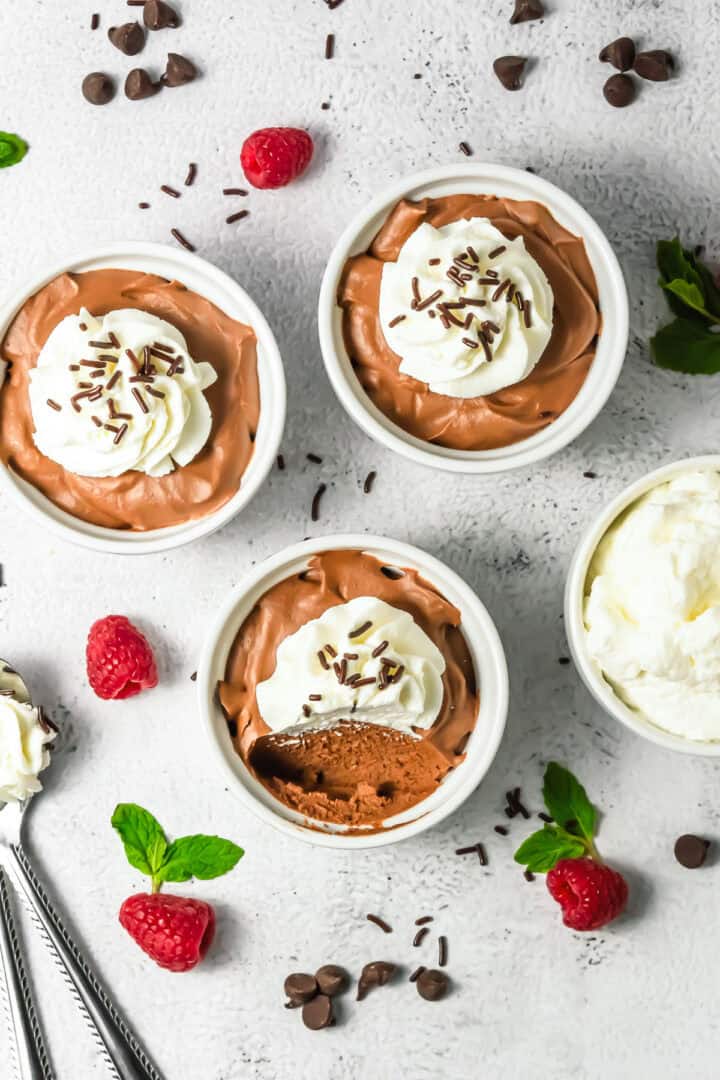 Three bowls of Chocolate Mousse topped with whipped cream and sprinkles, surrounded by fresh raspberries and mint, create an elegant dessert presentation.