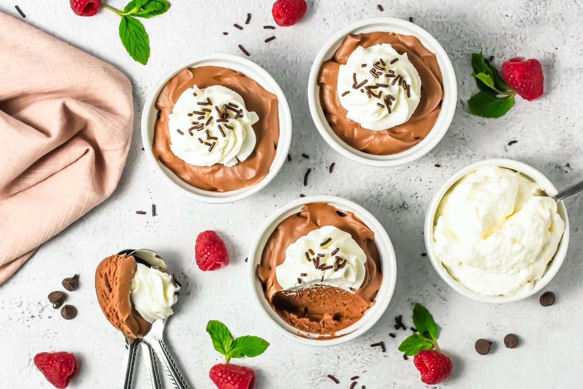 Three bowls of rich Chocolate Mousse with whipped cream and sprinkles, surrounded by fresh raspberries and mint leaves.