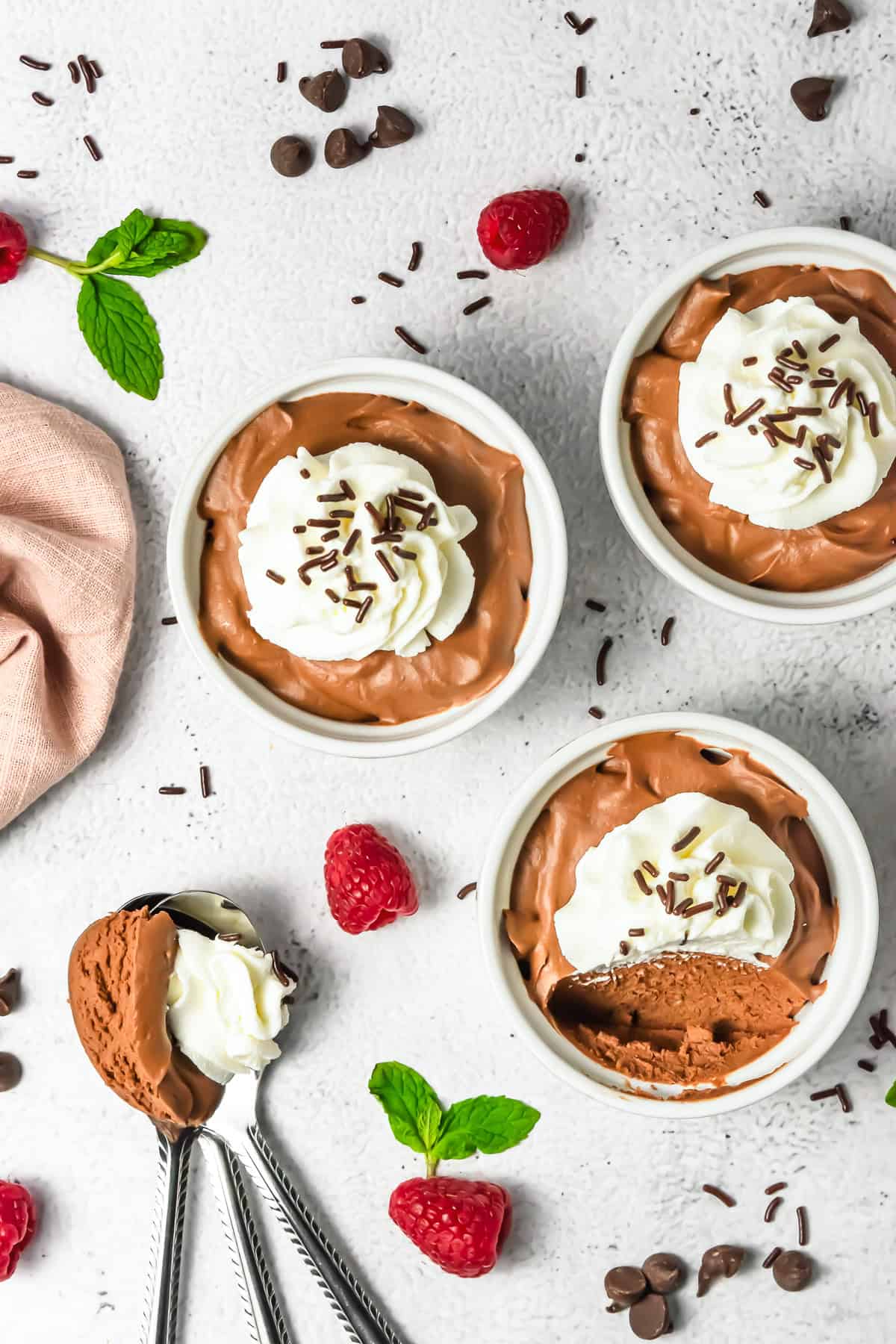 Three bowls of rich chocolate mousse topped with whipped cream and sprinkles, served alongside fresh raspberries and mint leaves.