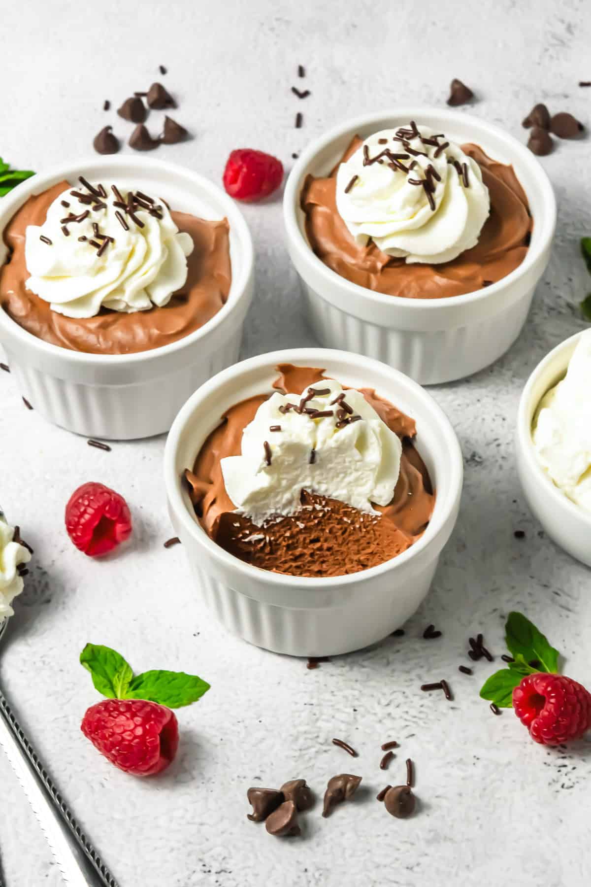 Four ramekins of rich Chocolate Mousse topped with whipped cream, garnished with fresh raspberries and chocolate chips nearby.