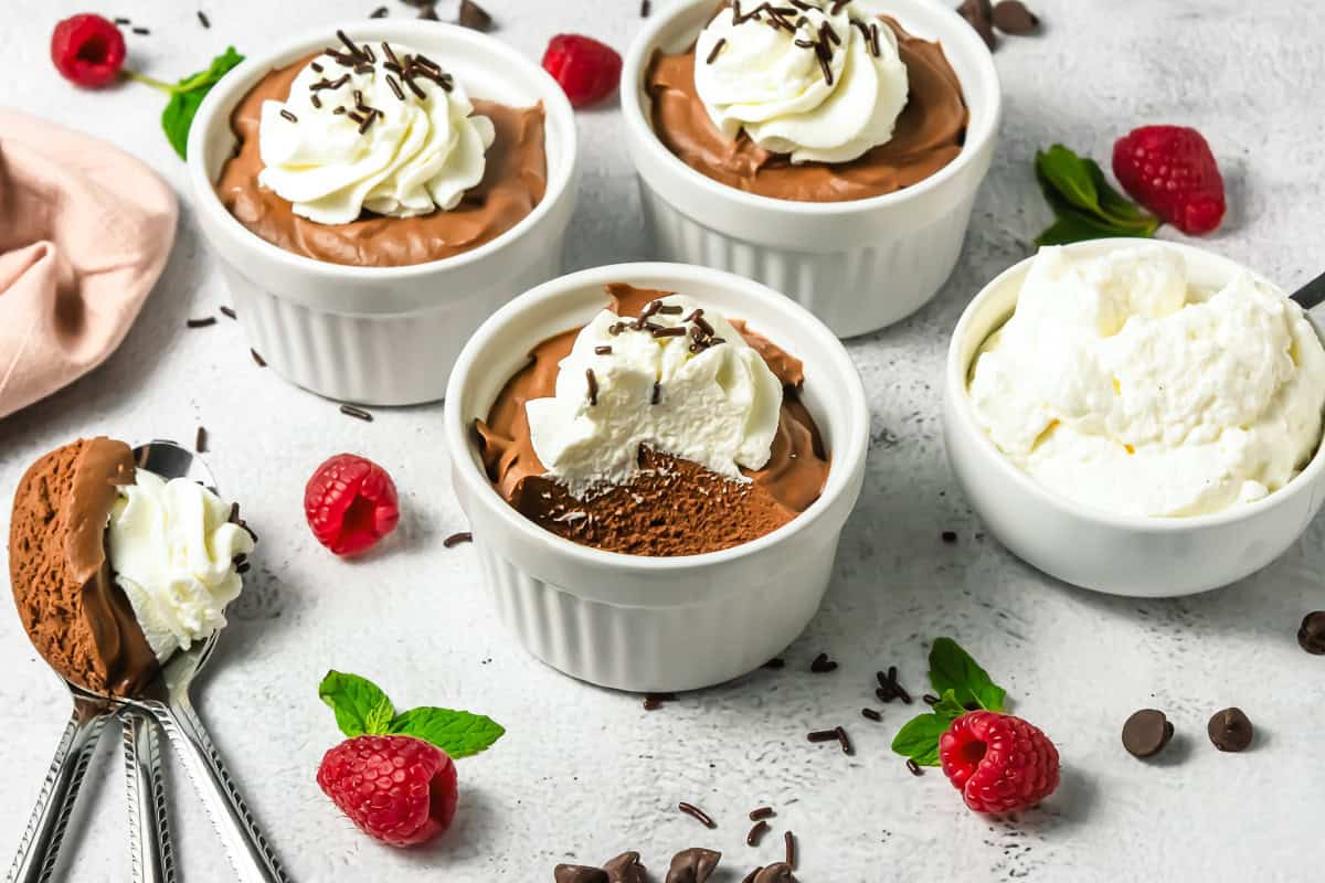 Three ramekins of rich chocolate mousse with whipped cream, surrounded by fresh raspberries, mint, and chocolate chips.