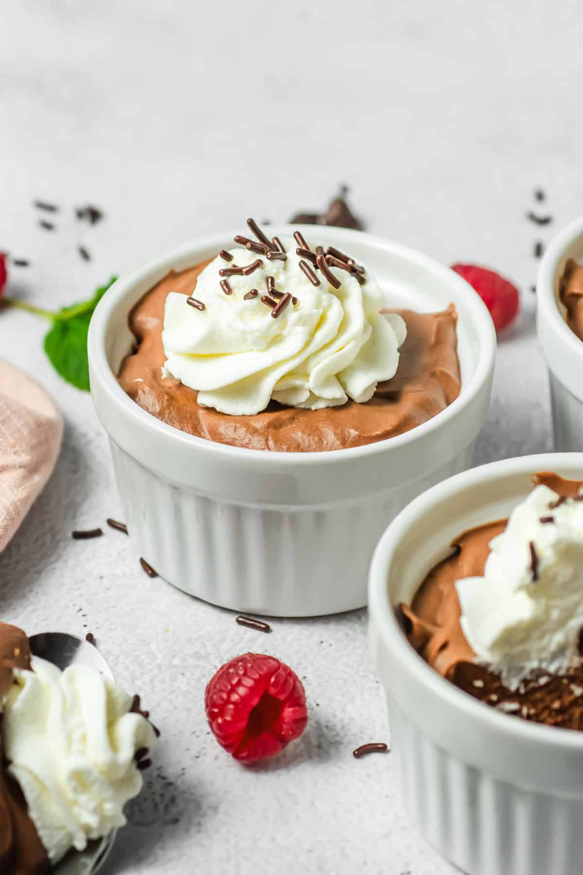 Chocolate Mousse in a white ramekin, topped with whipped cream and chocolate sprinkles, with fresh raspberries nearby.