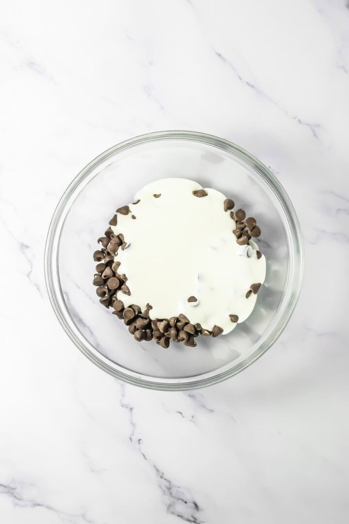 A glass bowl with chocolate chips and cream on a white marble surface, perfect for making rich Chocolate Mousse.