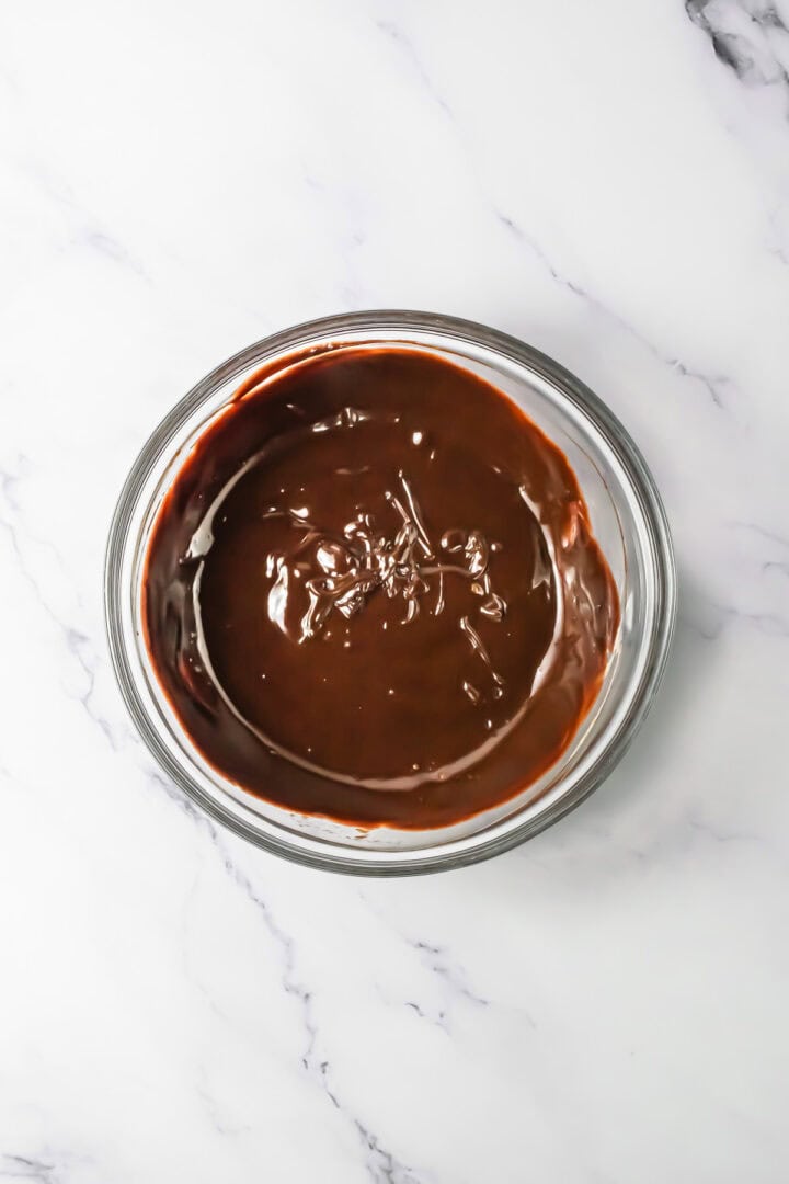 A glass bowl filled with melted chocolate, perfect for whipping up a decadent chocolate mousse, sits on a white marble surface.