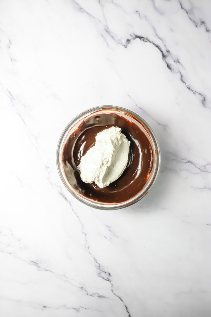 A glass bowl with rich chocolate mousse and a dollop of whipped cream sits on a white marble surface.