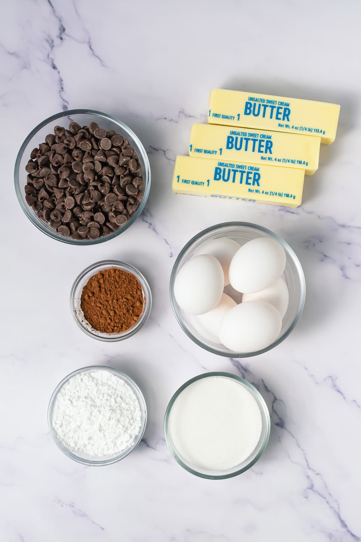 Bowls of chocolate chips, cocoa powder, powdered sugar, and eggs, plus butter sticks on a white marble surface.