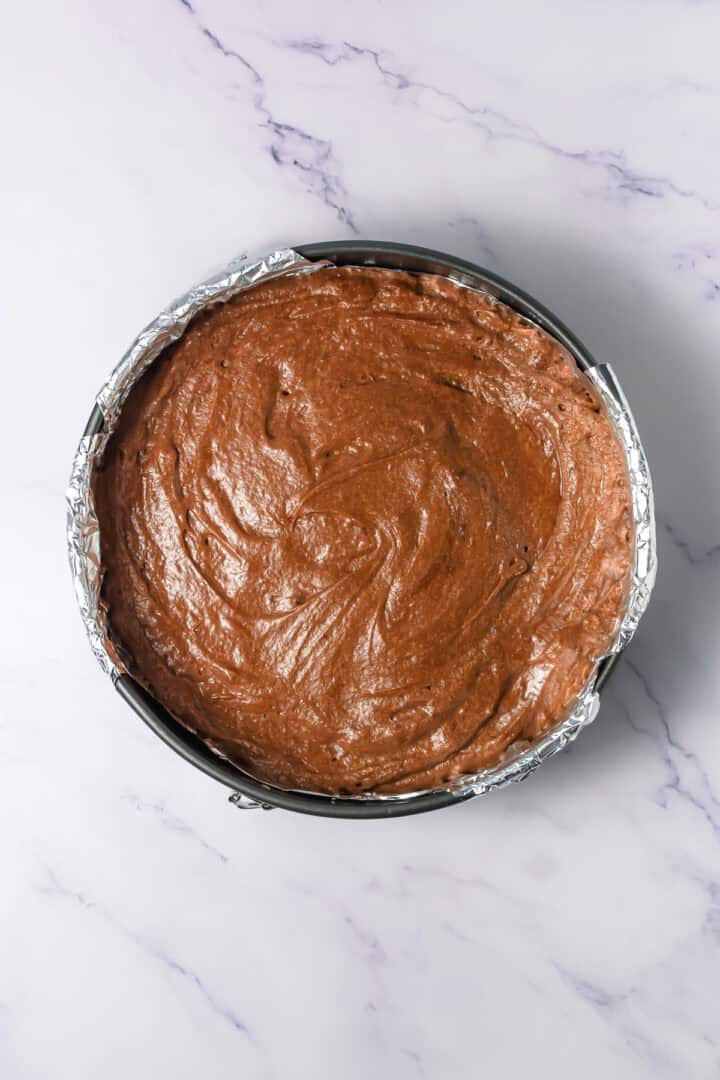 Round cake pan filled with chocolate batter on a marble surface, lined with foil.