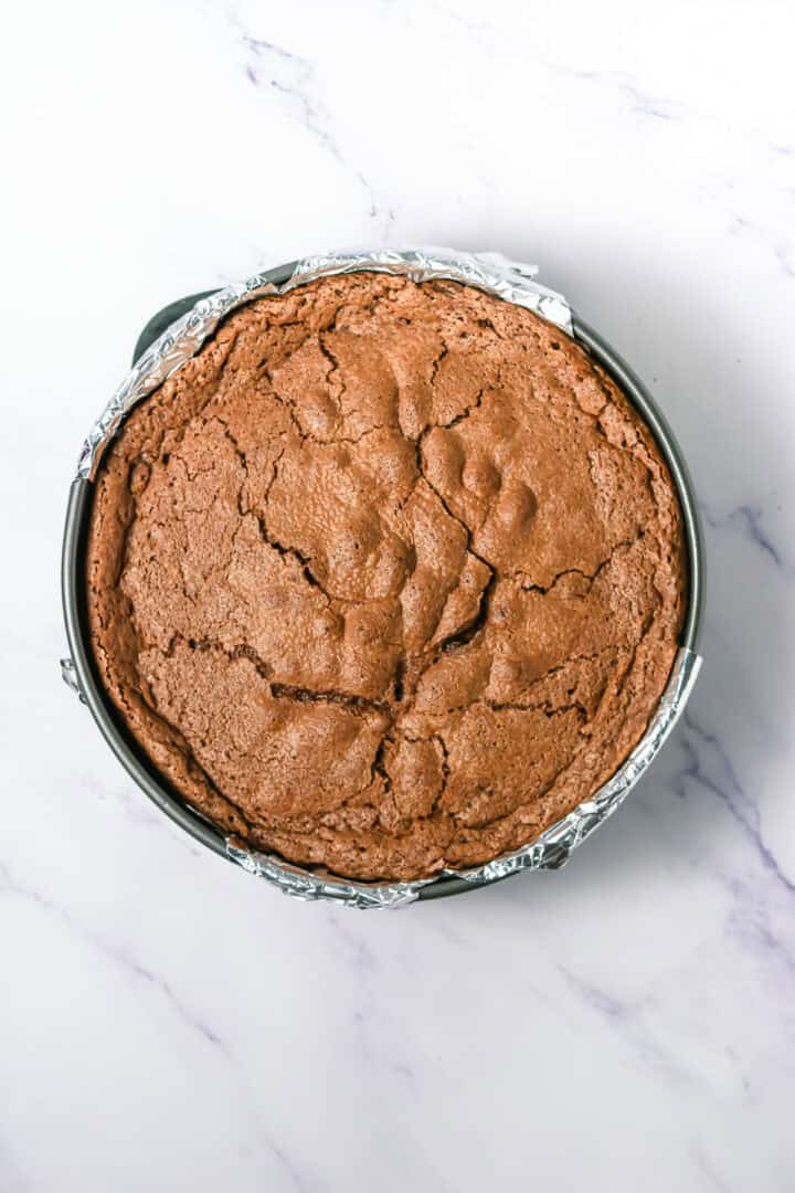 A round, cracked chocolate cake in a foil-lined baking pan on a marble surface.