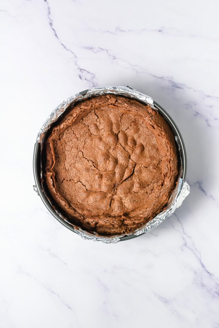 A round, cracked chocolate cake in a foil-lined springform pan on a marble surface.