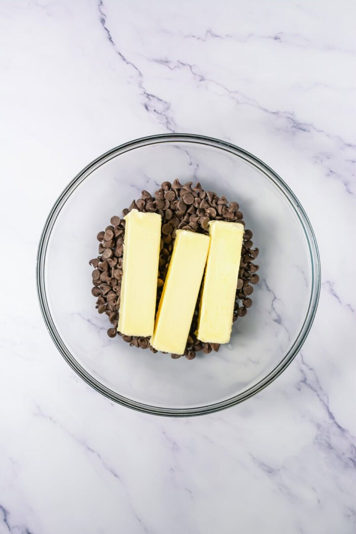 Three sticks of butter on top of chocolate chips in a clear glass bowl on a marble surface.