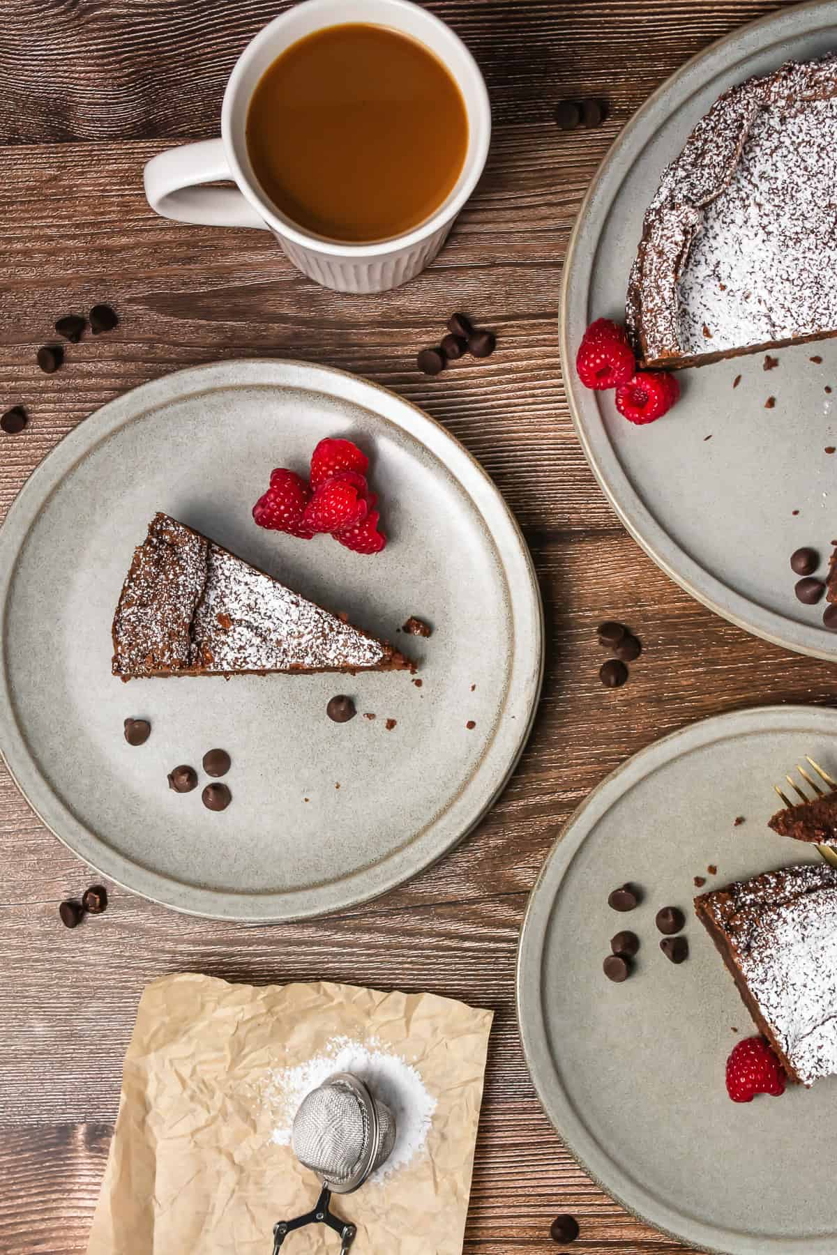 A slice of chocolate cake with raspberries and powdered sugar beside a cup of coffee on a wooden table.