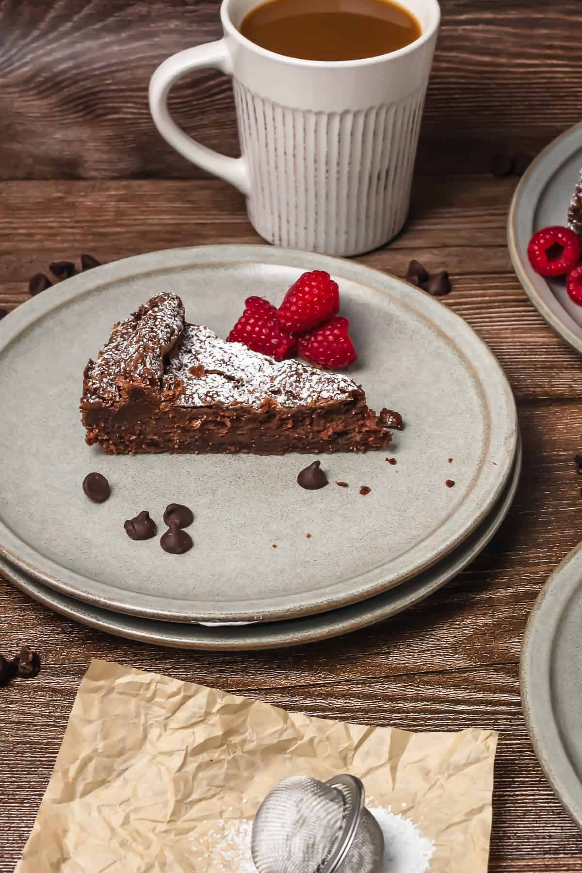 A slice of chocolate cake with raspberries and chocolate chips on a plate, next to a cup of coffee.