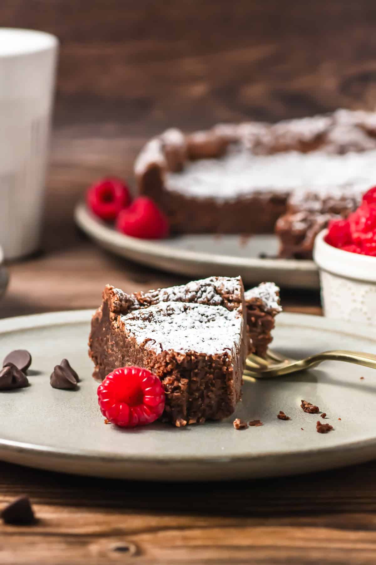 A slice of chocolate cake with powdered sugar, raspberries, and chocolate chips on a plate.