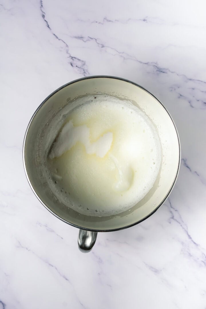 Metal mixing bowl with foamy, partially whisked egg whites on a marble surface.