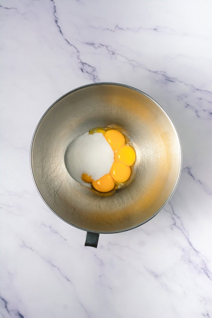 A metal bowl with egg yolks and sugar on a white marble surface, viewed from above.