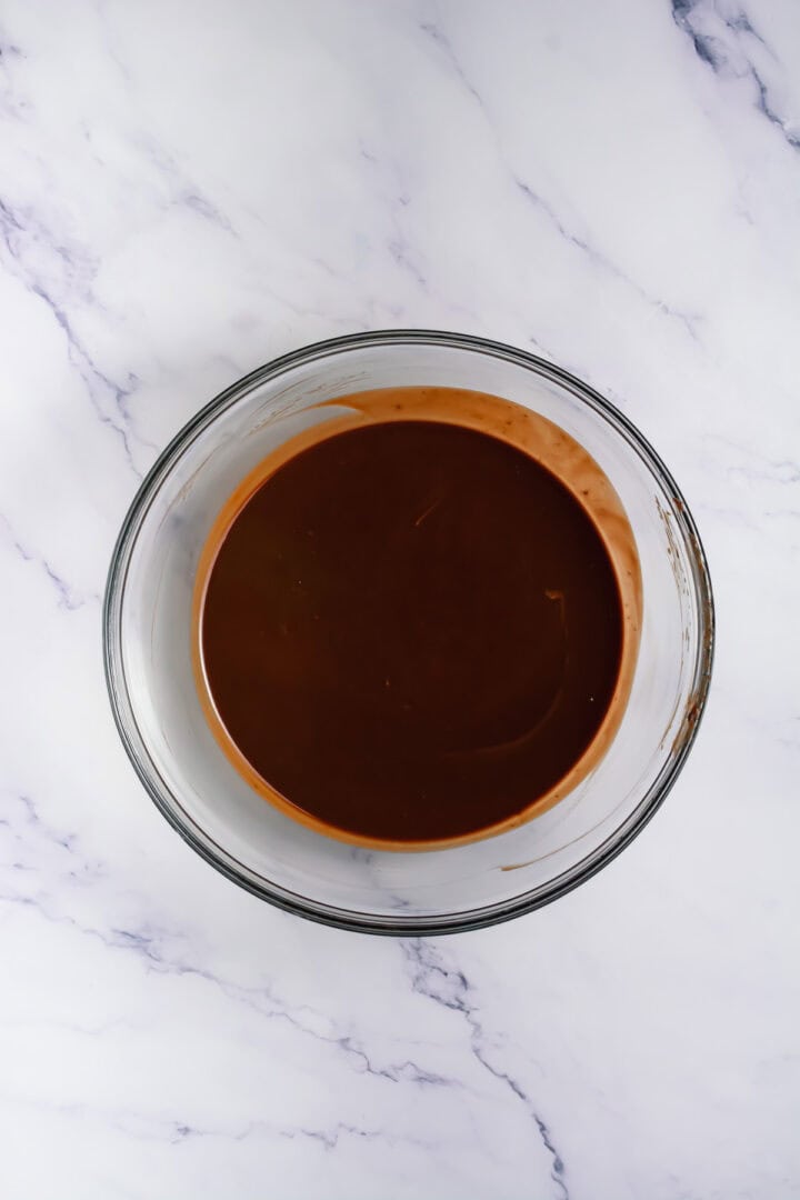 A glass bowl filled with smooth, melted chocolate on a white marble surface.