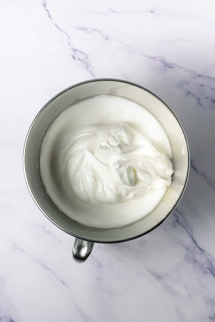A metal mixing bowl with glossy, whipped egg whites forming stiff peaks on a marble surface.