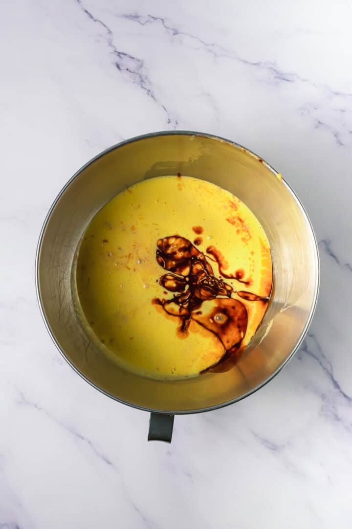 Metal mixing bowl with yellow batter and a splash of dark vanilla extract on a white marble surface.