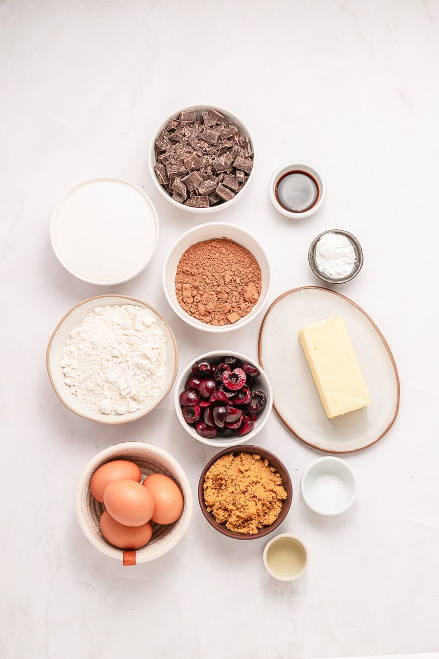 Bowls of chocolate chunks, cocoa, sugar, flour, eggs, cherries, butter, baking powder, and vanilla on a white surface.
