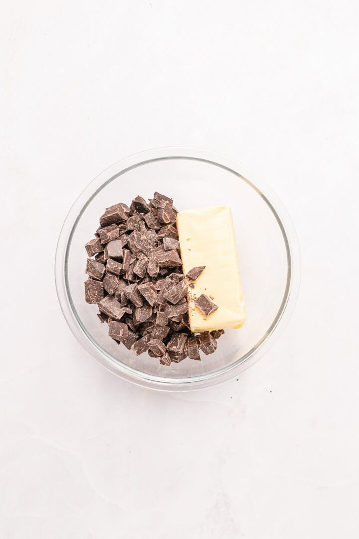 A glass bowl with chopped dark chocolate and a stick of butter on a white surface.