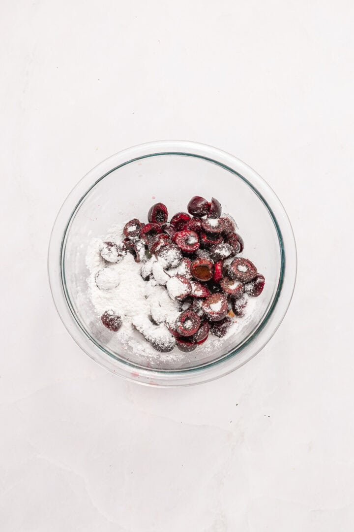 Glass bowl with halved cherries and white flour on a white marble surface.