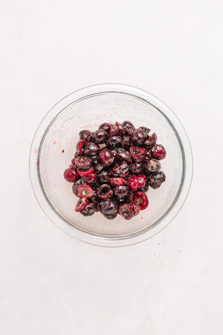 A glass bowl filled with halved, pitted dark cherries on a white surface.