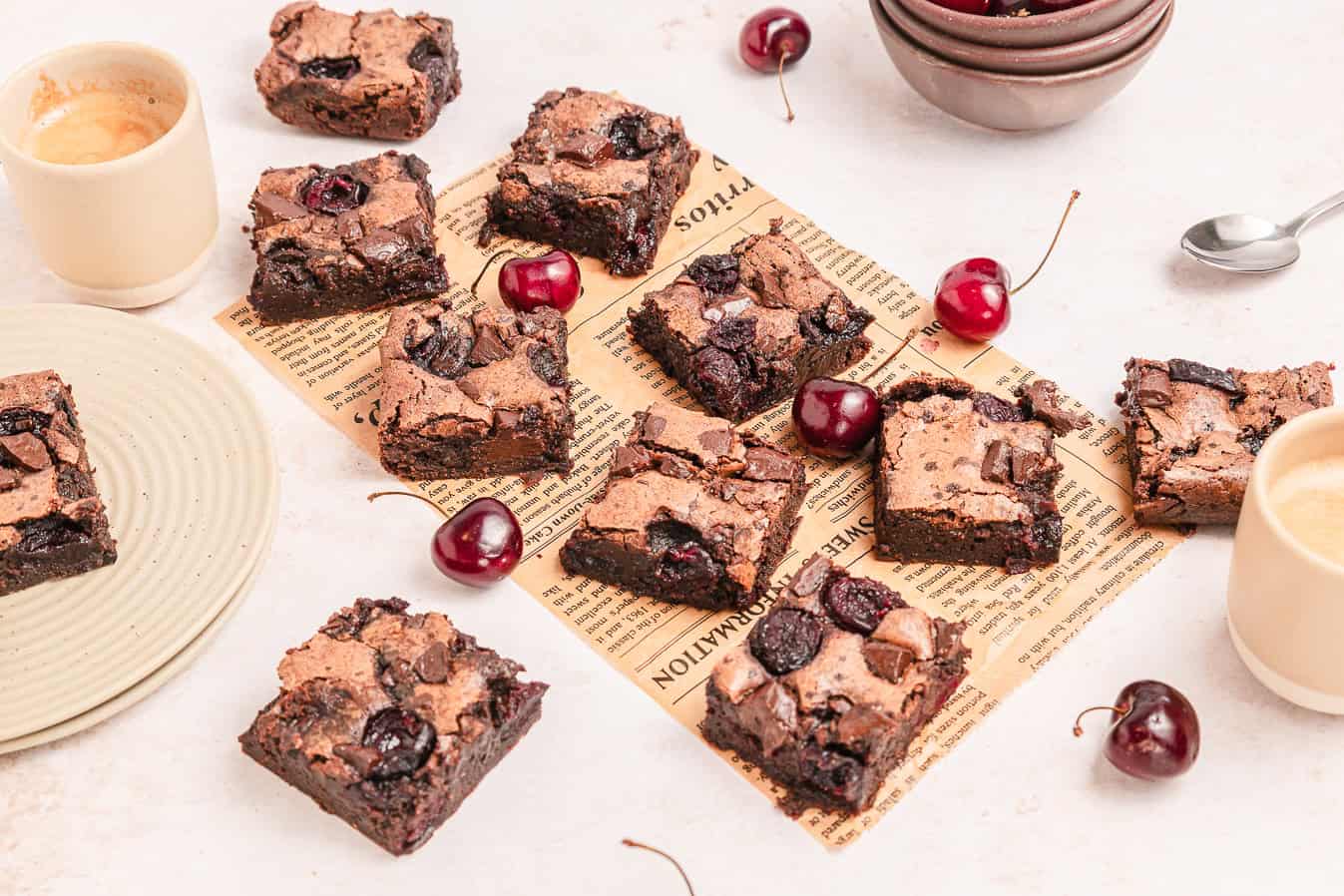Chocolate cherry brownies on newspaper, surrounded by fresh cherries, coffee cups, and dessert plates.