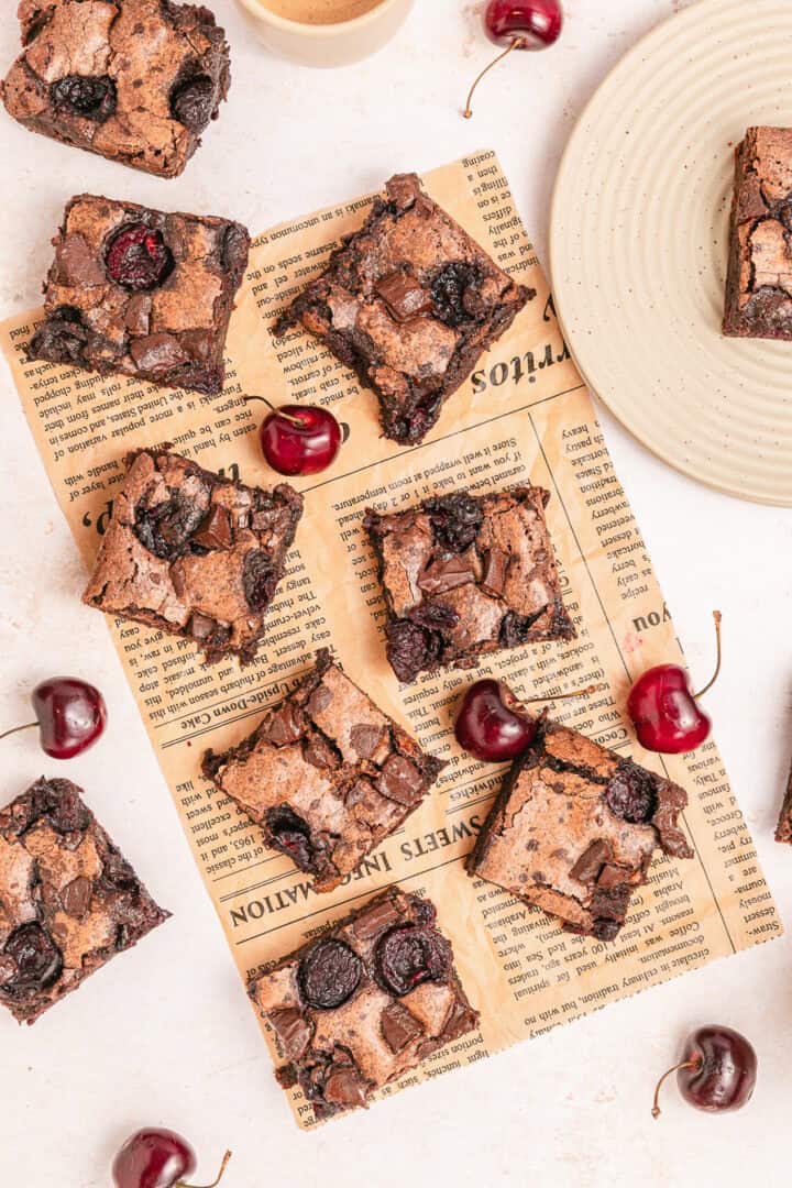 Brownie squares with cherries on and around them, displayed on a piece of newspaper and a white plate.