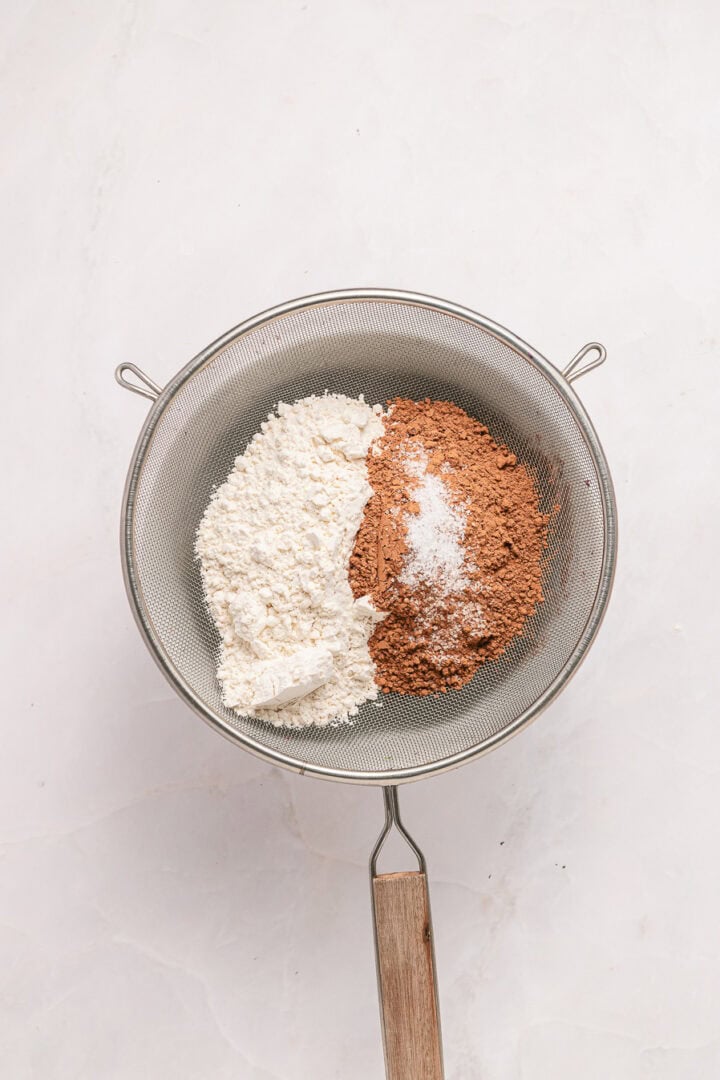 A sieve with flour, cocoa powder, and salt inside, resting on a light-colored surface.