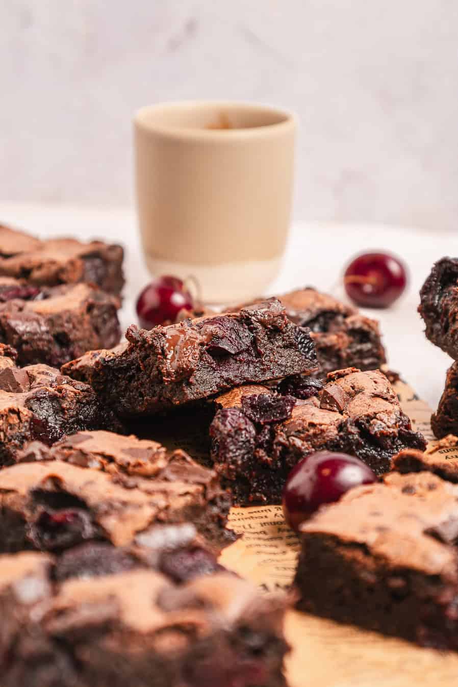 Fudgy chocolate brownies with cherries, some whole cherries, and a cup in the background.