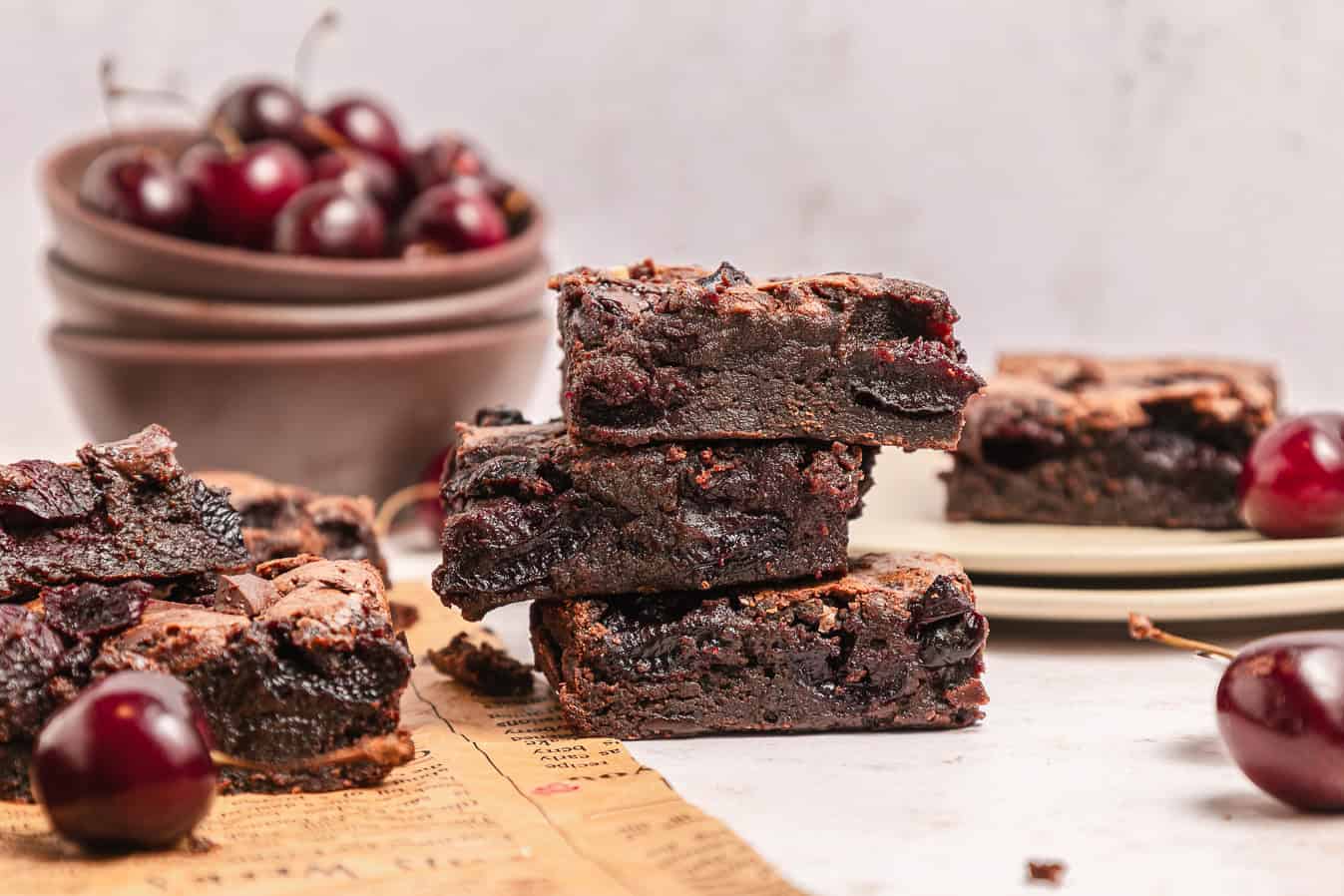 Stacked cherry brownies with fresh cherries and bowls in the background on a light surface.
