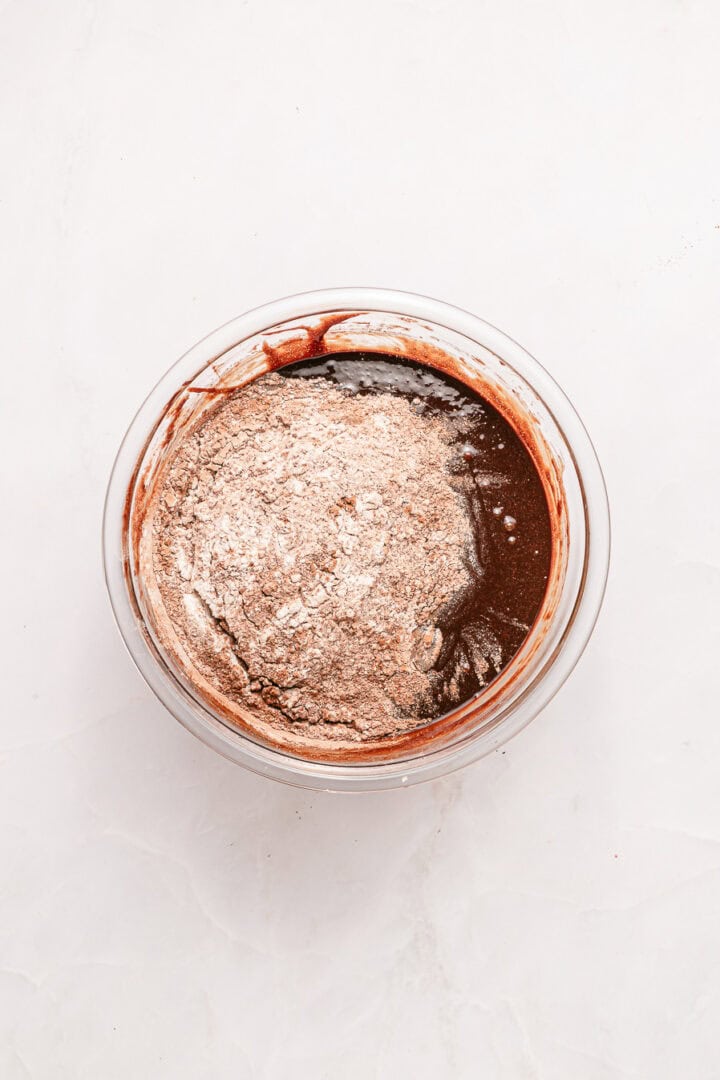 A glass bowl with chocolate batter and dry flour mixture on a white surface.