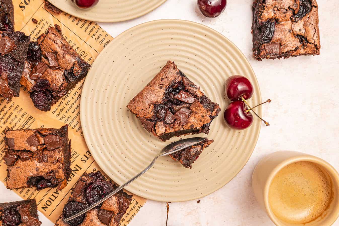 A plate with a chocolate brownie, two cherries, a fork, coffee cup, and more brownies on a light background.