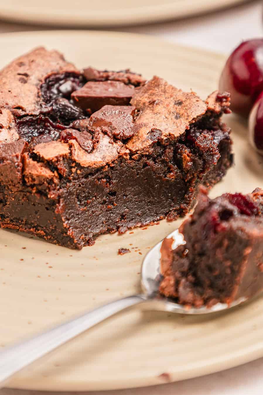 A close-up of a fudgy chocolate cherry brownie with a piece on a spoon, served on a beige plate.