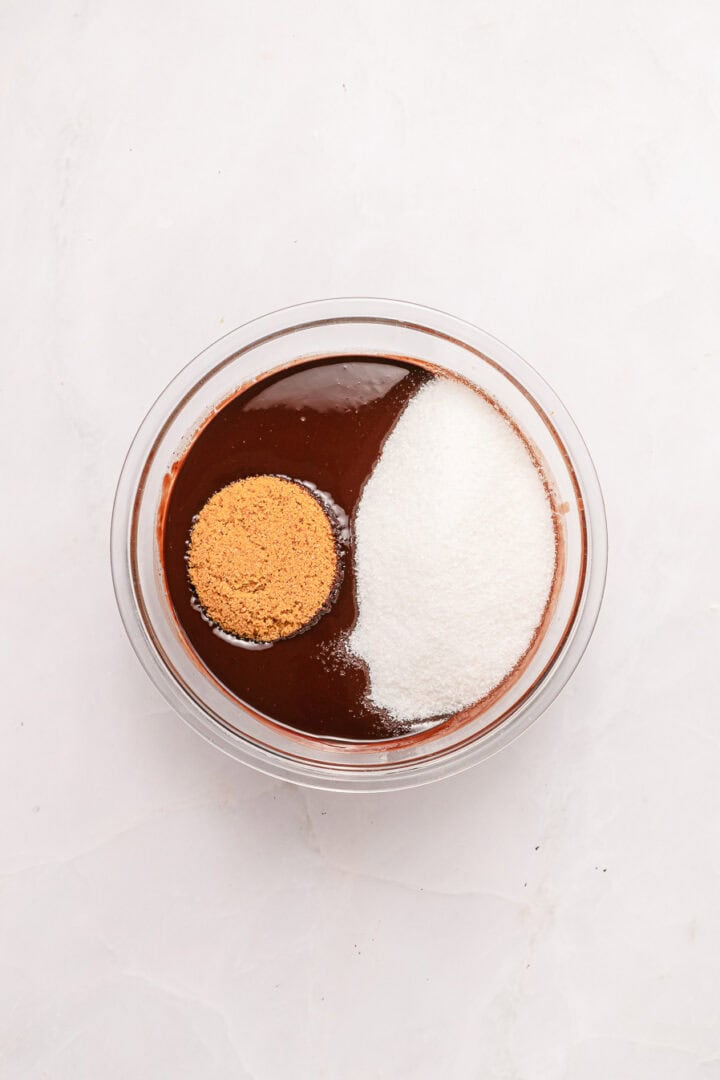 A glass bowl with cocoa, granulated sugar, and brown sugar on a white surface, viewed from above.