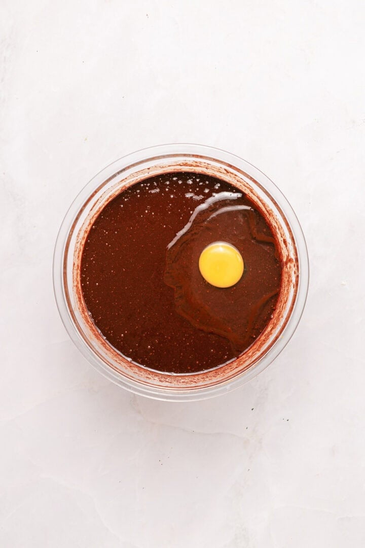 A glass bowl of chocolate batter with a raw egg on top, viewed from above on a white surface.