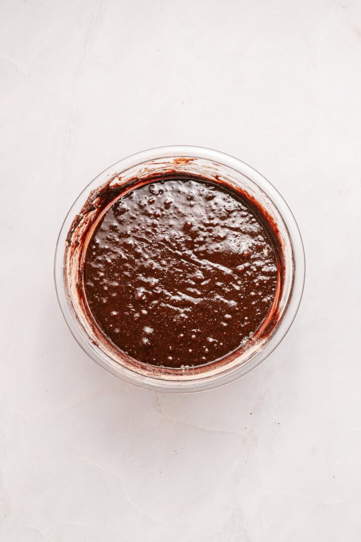 Glass bowl filled with chocolate cake batter on a white marble surface.