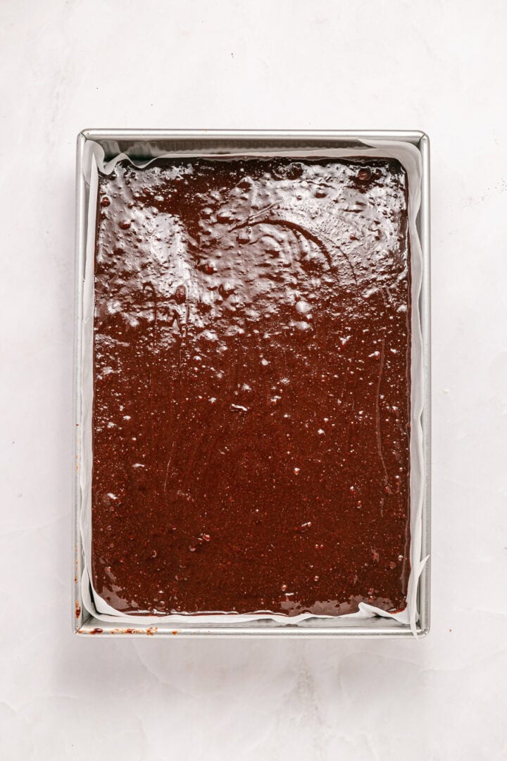 Square baking pan filled with chocolate brownie batter on a white surface, ready to be baked.