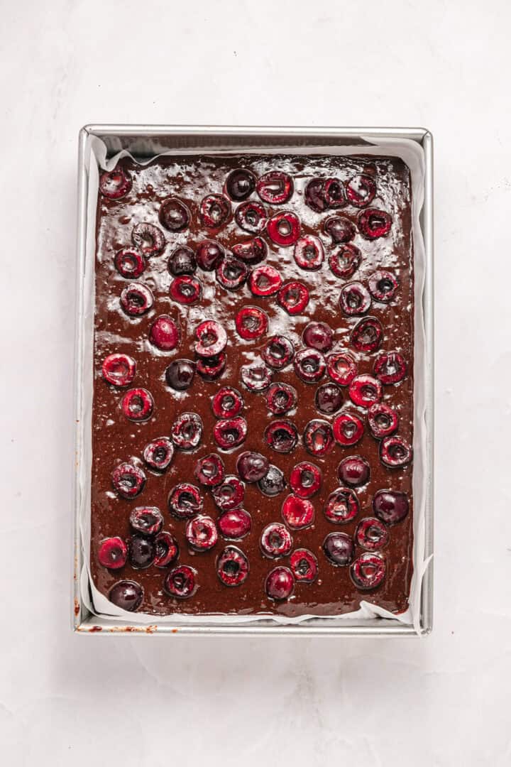 A rectangular pan of chocolate batter topped with halved cherries on a white surface.