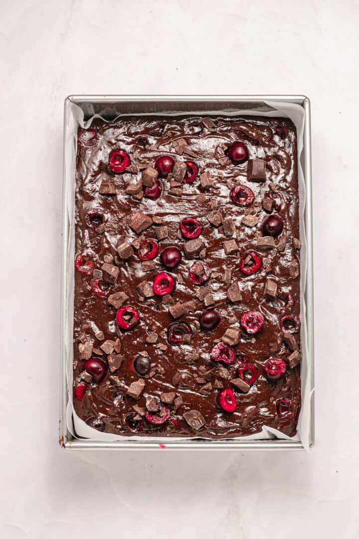 A tray of unbaked chocolate brownie batter topped with cherries and chocolate chunks on a marble surface.