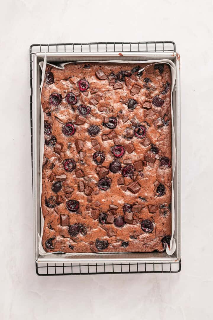 A baked chocolate cherry cake in a parchment-lined rectangular pan on a cooling rack.