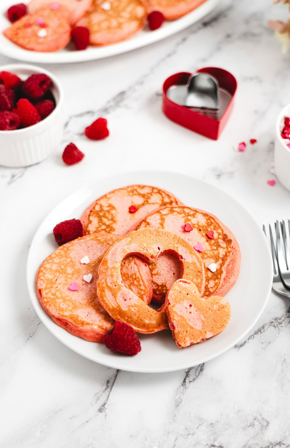 A plate of Raspberry Valentine’s Pancakes, shaped like hearts and topped with raspberries and heart-shaped sprinkles, sits beautifully on a white table.
