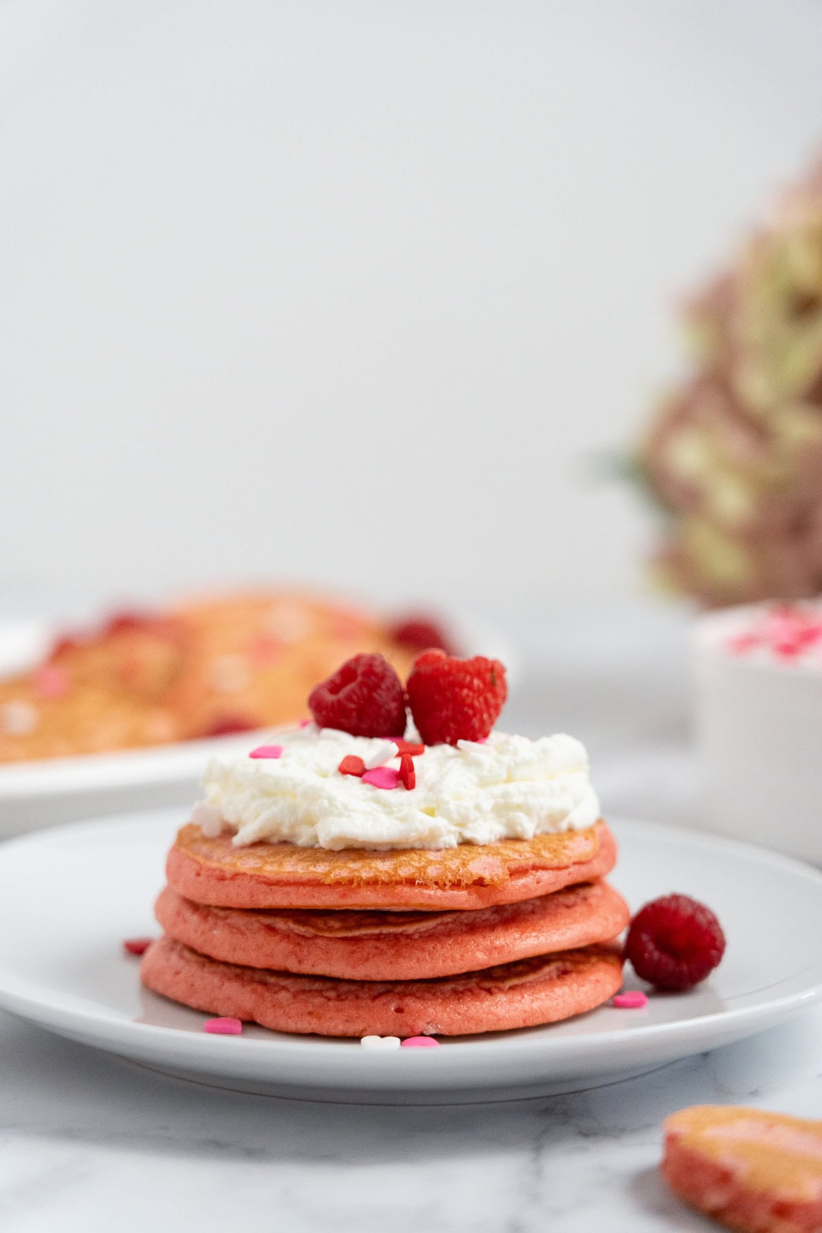 Stack of Raspberry Valentine’s Pancakes topped with whipped cream, raspberries, and heart sprinkles on a white plate.
