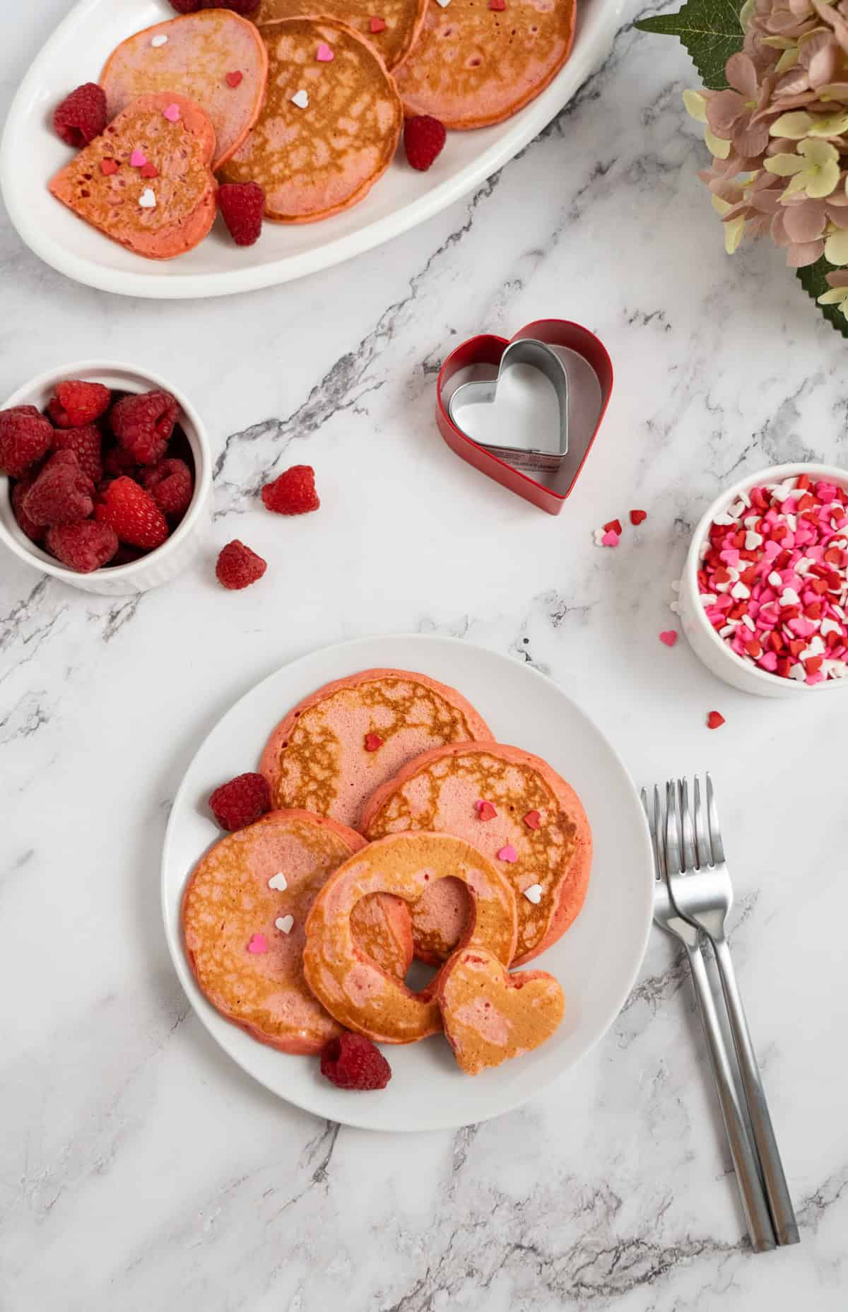 Raspberry Valentine’s Pancakes featuring pink pancakes with heart-shaped cutouts, fresh raspberries, and heart sprinkles on a marble table.