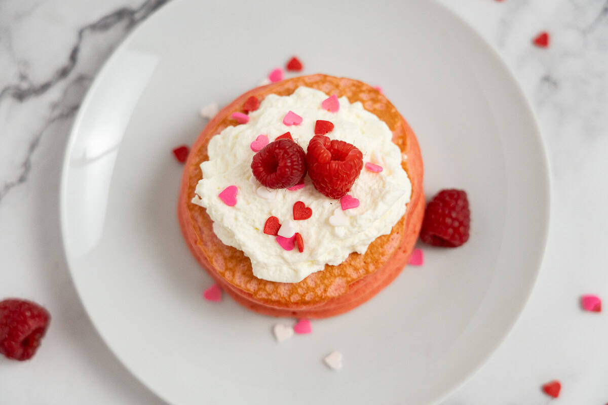 A pink Raspberry Valentine’s Pancake topped with whipped cream, fresh raspberries, and heart-shaped sprinkles on a white plate.