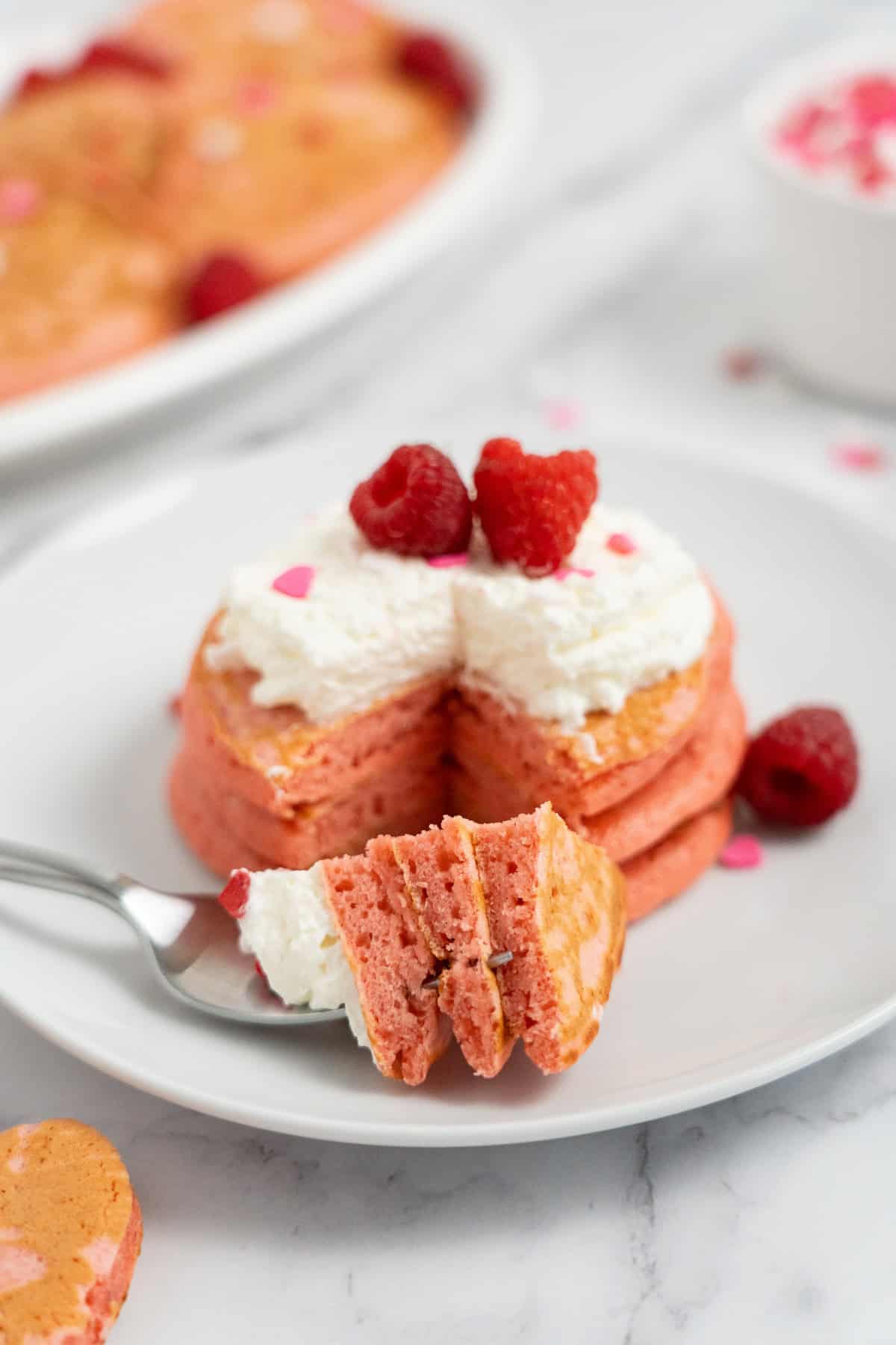 Stack of pink Raspberry Valentine’s Pancakes topped with whipped cream and raspberries; a fork holds a bite-sized piece.