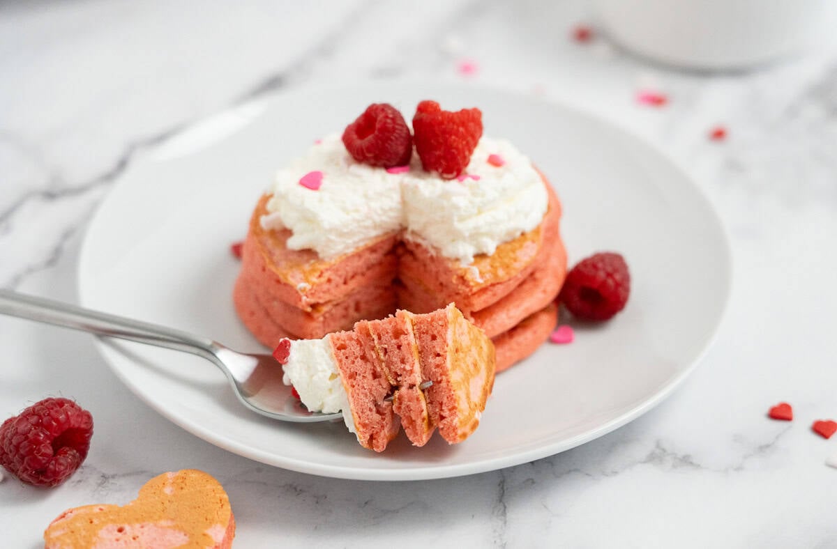 A fork holds a bite of Raspberry Valentine’s Pancakes topped with whipped cream and raspberries on a white plate.